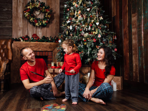 Two parents and one child wearing matching holiday pajamas and sitting around a Christmas tree