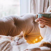 Woman sitting on couch with fuzzy socks, large blanket, a mug, and a cat on her lap